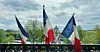 French flags waving above a bridge railing with Paris buildings in the distance.