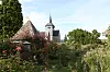Church tower above a flower-filled garden with a small stone-roofed structure and greenery.