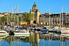 Panoramic harbour view with sailboats, waterfront buildings and a city skyline reflected in calm water.