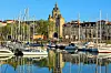 Panoramic harbour view with sailboats, waterfront buildings and a city skyline reflected in calm water.