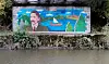 Colourful mural on a wall beside a river, showing a portrait, boats and landscape imagery.
