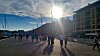 Crowds walk along a sunny waterfront promenade in Marseille with long shadows and boats in the background.