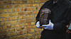 Undertaker in dark clothing holding a metal urn beside a brick wall.