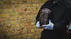Undertaker in dark clothing holding a metal urn beside a brick wall.