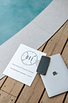 Brochure, smartphone and MacBook on a wooden deck beside a swimming pool.