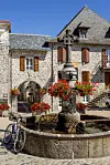 Stone fountain with red flowers in a cobbled village square next to a bicycle and old stone buildings.