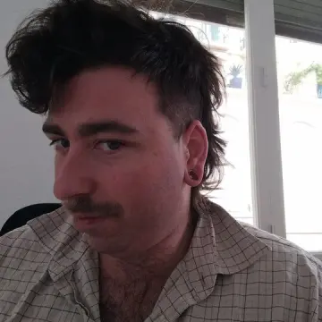 Man with dark tousled hair and a checked shirt sits indoors beside a window.