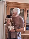 An older man holding a cat indoors beside a wooden cabinet.