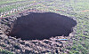 Large sinkhole in a green cultivated field with exposed dark soil and debris.