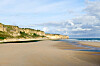 Wide sandy beach beside cliffs and calm sea at Omaha Beach under a partly cloudy sky.