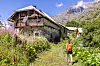 Person walking along a path beside a rustic farmhouse in a mountainous rural landscape.