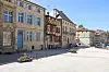 Sunlit town square in Bar-le-Duc, France, with historic buildings, flower planters and parked cars.