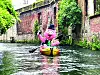 A pink mascot paddles a yellow kayak in a canal flanked by brick walls and plants.