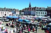 Crowded outdoor market in a town square with stalls and historic buildings under a blue sky.