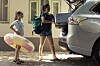 Mother and daughter loading a suitcase into the back of a silver car outside a house.