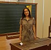 Woman standing in a classroom beside a blackboard and wooden desk.