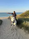 A rider on a white horse points towards the sea on a sandy beach.