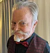 An older man in a red bow tie and patterned waistcoat poses indoors beside curtains.