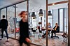 People work and move through a modern glass-walled office with meeting tables and pendant lights.