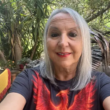 Silver-haired woman smiling for a selfie in a lush garden, wearing a black T-shirt with a red feather design.