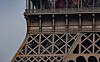 Close-up of the Eiffel Tower’s iron lattice, arches, columns and lamps against a pale sky.