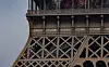 Close-up of the Eiffel Tower’s iron lattice, arches, columns and lamps against a pale sky.