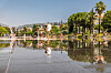 People walk beside a wet street reflecting palm trees and buildings on a sunny day.