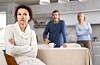 Woman with folded arms sits apart as a man and mature woman stand behind her in a tense home setting.