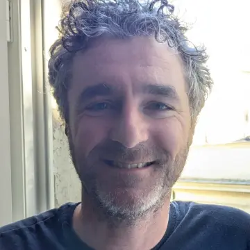 Bearded man with curly hair smiling beside a bright window