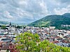 Panoramic city centre with red rooftops, church towers and green mountains under a cloudy sky