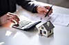 Hands using a calculator beside a miniature house and property paperwork