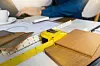 Timber samples, plans, a ruler and yellow tape measure on a table beside a seated person.