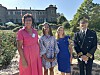 Three women and a uniformed officer pose beside a rose garden outside a historic stone building.