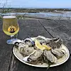 Plate of oysters with lemon beside a jar of golden drink on a seaside table.