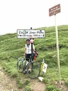 Cyclist with a road bike beside the Col de Joux Plane sign in a grassy mountain landscape