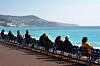 People sit in blue chairs along Nice seafront overlooking the Mediterranean.