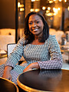 Robin Davis smiles at a café table in a warmly lit Parisian interior, wearing a black-and-white patterned dress.