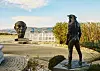 Bronze statue on a seaside promenade with a second sculpture and coastal landscape behind it