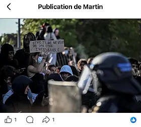 Masked protesters hold a handwritten placard as a helmeted police officer stands in the foreground.