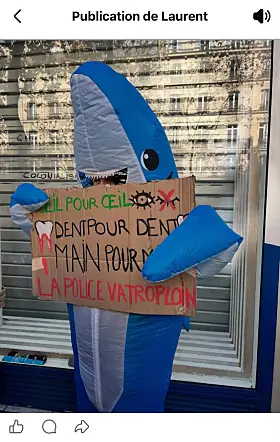 Blue inflatable figure holding a handwritten protest placard outside a shuttered shop