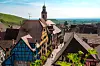 Historic half-timbered village street with a church tower, red-roofed houses and green hills beyond.